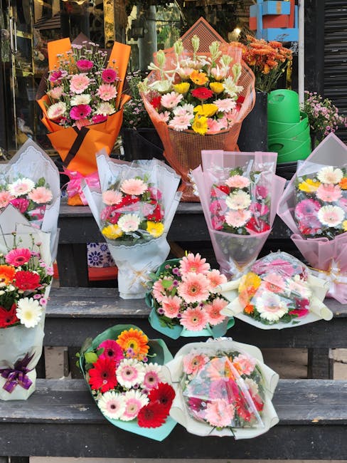 A display of various fresh floral bouquets arranged on a wooden outdoor table, featuring a mix of pink, white, yellow, and red daisies, chrysanthemums, and gerbera daisies. The bouquets are wrapped in