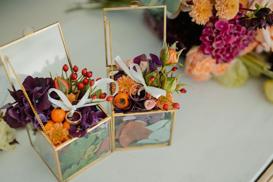 A pair of floral arrangements displayed inside small, open glass and gold-framed boxes on a white surface. Each arrangement features a mix of fresh flowers, including deep purple calla lilies, orange 