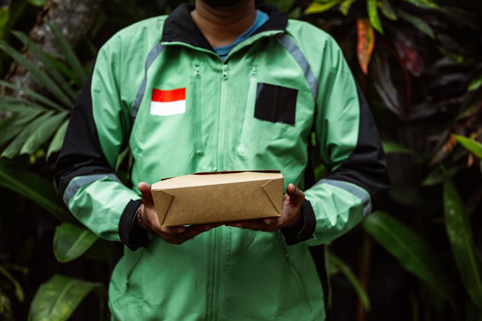 A delivery person wearing a green jacket with black sleeves, a black patch, and a small Indonesian flag patch on the chest, is holding a rectangular brown cardboard parcel in both hands. The backgroun