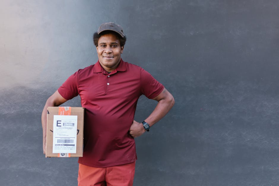 A male delivery person with dark skin, smiling and wearing a maroon polo shirt, maroon pants, and a dark baseball cap, holding a cardboard parcel with a barcode label and shipping information. He stan